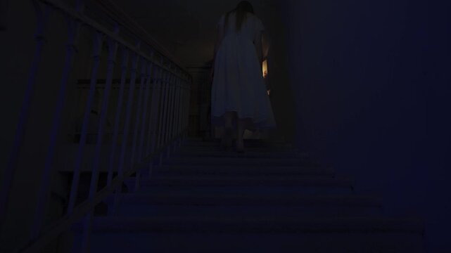 Creepy ghost woman in white dress and scary makeup walking up alone on staircase in dark haunted house. Halloween horror advertisement concept.