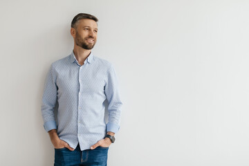 A man with a beard stands casually, smiling while leaning against a plain wall. He is dressed in a...