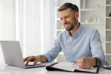 The man focuses intently on his laptop, taking notes in a notebook. Sunlight floods the modern space, highlighting his productive atmosphere and diligent effort.