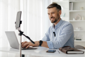 Obraz premium A man is seated at a bright desk, focused on his laptop as he prepares for a video call. He appears engaged and ready to communicate professionally.
