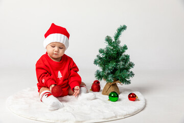 Top view portrait of cute little newborn baby girl, wearing red Santa hat holds Christmas balls near Christmas tree on white background. Christmas and New Year concept. High quality photo, copy space