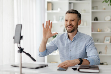 A man with a friendly smile gestures while interacting during a virtual meeting from his home office, surrounded by books and decorative items.