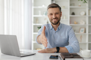 A man in a light blue shirt smiles as he reaches out for a handshake from a home office setting filled with natural light, indicating a friendly virtual meeting.