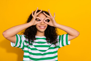 Photo of nice young girl fingers binoculars look empty space wear striped t-shirt isolated on yellow color background