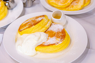 A plate of fluffy souffle pancakes served with a caramelized banana, powdered sugar, whipped cream, and a side of chocolate syrup, creating a decadent dessert