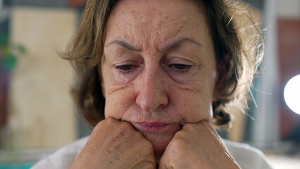 Elderly woman sitting at the table, resting her chin on hands, displaying a preoccupied and anxious...