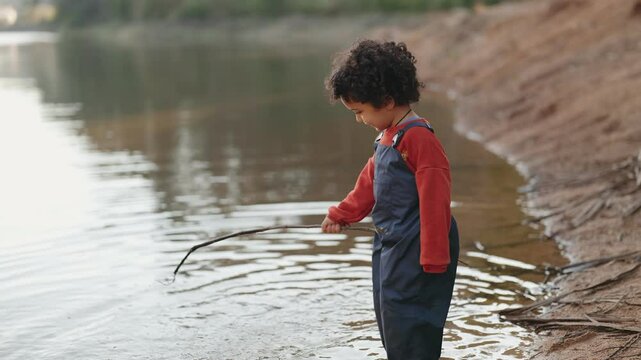 Curly-haired child in overalls plays with a stick in the water, creating splashes on a tranquil lakeside.