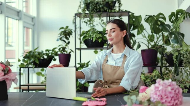 Confident young female entrepreneur and florist smiles at the camera in her plant and flower shop. She closes her laptop after a hard day of work running her small business.