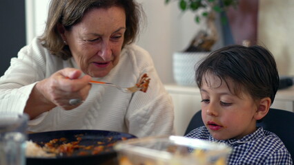 Grandmother gently feeding reluctant young boy at the table, experiencing a moment of care and...