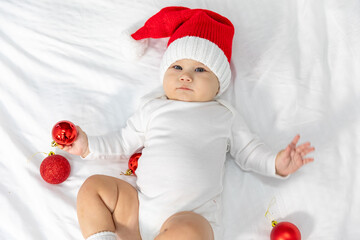 Close-up top view portrait of a newborn baby. Cute Caucasian baby 6-7 months old in red Santa hat and white suit costume lying on a cozy bed with Christmas ball in hand.