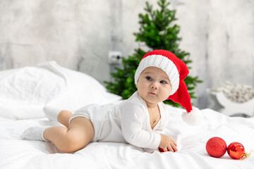 Close-up portrait of a newborn baby. Cute Caucasian baby 6-7 months old in red Santa hat and white suit costume lying on a cozy bed near a decorated Christmas tree. Merry Christmas 2025. Side view