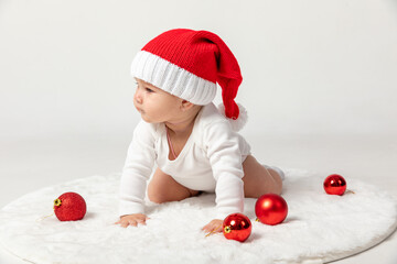 Portrait of newborn baby girl, wearing red Santa hat and white body suit on a white fur carpet on white background among Christmas balls. New Year holidays concept, copy space, high quality photo