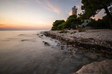 Beautiful sunset in a landscape on a rocky coast with a striking lighthouse and pine forest. View...
