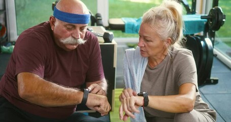 Senior retired couple checking results on their smartwatches after training at home gym, talking and sharing information, goals achieved, heart rate measurement, and training time - Powered by Adobe