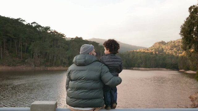 A tender moment as a father holds his child, both overlooking a calm lake surrounded by forests at dusk.
