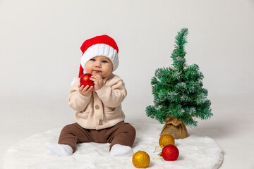 Cute Christmas little baby in a red Santa hat with Christmas balls and little Christmas tree on a white background. Christmas and New Year celebration