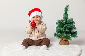 Cute Christmas little baby in a red Santa hat with Christmas balls and little Christmas tree on a white background. Christmas and New Year celebration