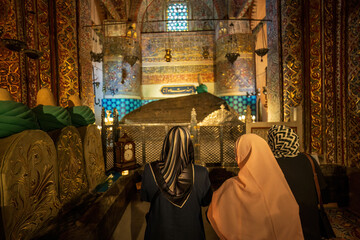 Three unidentified women wearing headscarves visit a shrine, kneeling in prayer before a decorated...