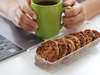 Businesswoman sitting behind a laptop, holding a coffee cup and getting ready to eat chocolate chip...