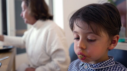 Young boy with food in his mouth looking distracted while sitting at the dining table. Unfocused...