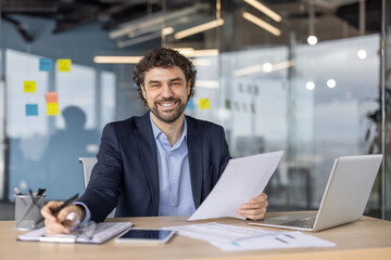 Confident businessman smiling in office holding document at desk with laptop and smartphone. Professional attire indicates leadership and success in vibrant workspace.