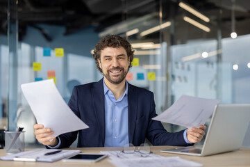 Obraz premium Smiling businessman in suit holding documents at desk with laptop and smartphone. Modern office setting with determined expression suggesting success, confidence, and professional achievement