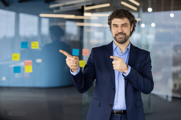 Businessman in office setting pointing at glass wall with sticky notes. Professional wearing blue suit projects confidence and positivity. Displays concept of teamwork