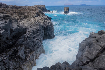 a cliff of black volcanic rock with a sea of turquoise water with waves lapping against the rocks, a picturesque rock rising out of the water