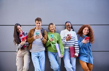 Group of smiling young multiracial friends leaning in grey wall using and looking colorful cell...