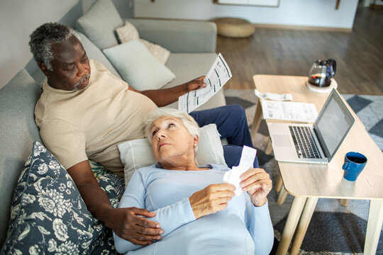 Diverse senior couple reading bills on the couch at home - Powered by Adobe