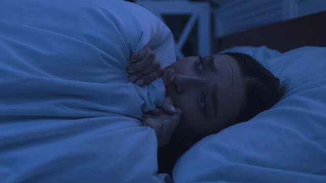 Worried girl lying awake in bed, covering face with blanket, peeking and looking to sides in fear