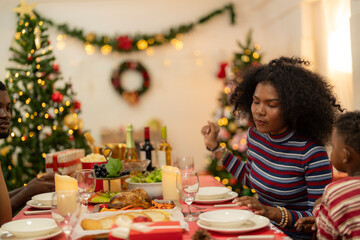 family moment during Christmas as they exchange gifts around festive dinner table. The table with Christmas-themed tableware, candles and delicious food. The room is adorned with holiday decorations.