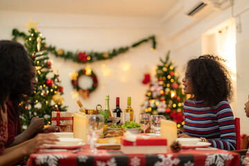 family moment during Christmas as they exchange gifts around festive dinner table. The table with Christmas-themed tableware, candles and delicious food. The room is adorned with holiday decorations.