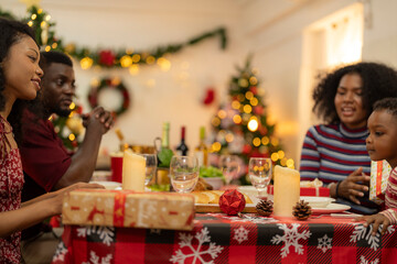 family moment during Christmas as they exchange gifts around festive dinner table. The table with Christmas-themed tableware, candles and delicious food. The room is adorned with holiday decorations.