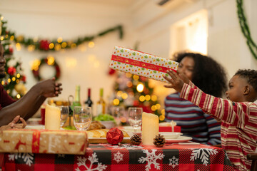 family moment during Christmas as they exchange gifts around festive dinner table. The table with Christmas-themed tableware, candles and delicious food. The room is adorned with holiday decorations.