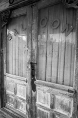 A pair of weathered, historic, abandoned wooden saloon doors with the word "SALOON" in capital letters across the glass panels are visible, located in a ghost town in Grove, Texas.