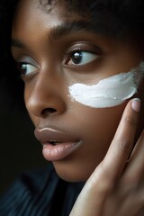 African American woman gently applies white facial cream to face in dark studio setting, dark hair, black top. Highlights intimate atmosphere of beauty studio, perfect for promotional.