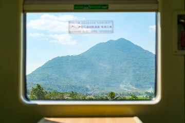 A breathtaking view of a mountain framed by a train window on a West Java route, with clear blue skies and lush greenery, capturing the beauty of Indonesia&rsquo;s natural landscapes