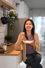 Pretty young asian woman eating a healthy breakfast at kitchen counter