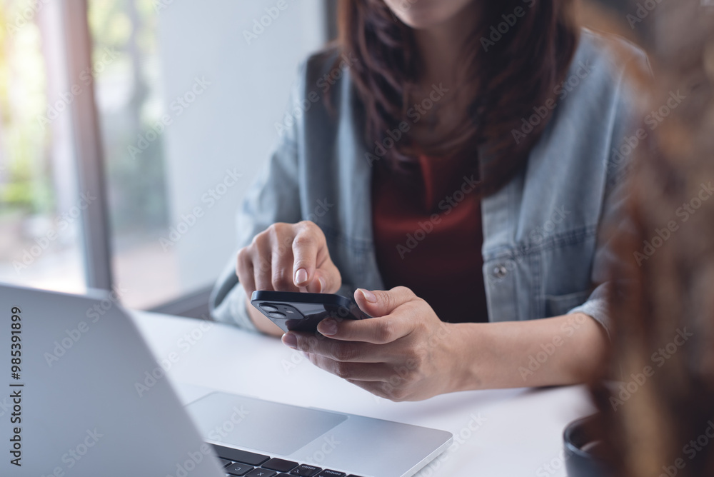 Poster Asian business woman using mobile phone with laptop computer on table. Woman surfing the internet, searching the information on smartphone during working on laptop computer at home office