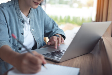 Woman working on laptop computer, taking note on notebook on table, at coffee shop, close up. Student studying online, searching the information and taking note on notepad,  e-learning