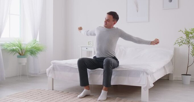 Happy man wakes up in the morning and stretching while sitting on the bed at home. He enjoys a relaxed moment after sleep, feeling refreshed and content as he starts day with a morning routine.