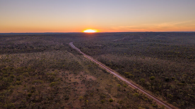 A long, straight road cuts through a vast, dense forest under the sunset sky.