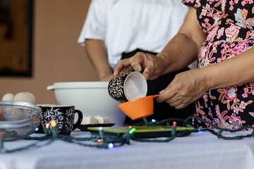 Primer plano de unas manos midiendo harina en una taza para una receta de repostería navideña. Sobre la mesa hay luces festivas e ingredientes como huevos.