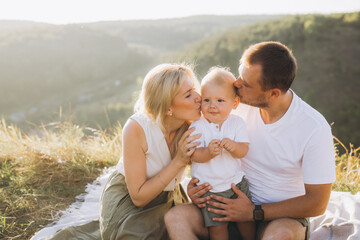 Happy Family Moment Outdoors With Parents Kissing Baby On Picnic Blanket In Sunshine