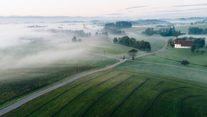 Aerial panoramic view of misty morning in rural Allgäu countryside with green fields, farms, and...