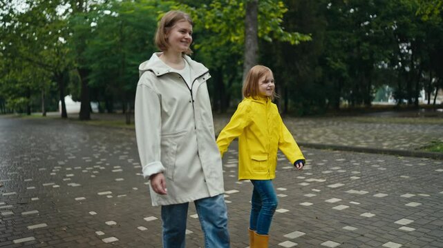 A happy blonde woman in a white jacket walks hand in hand with her teenage daughter in a yellow jacket walking in the park after the rain