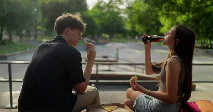 Happy couple of teenage classmates eating pizza and drinking soda while picnic and relaxing at skatepark in summer