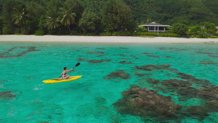 Aerial view of a tourist paddling on a yellow kayak in a tropical lagoon with turquoise water. The resort is surrounded by lush vegetation