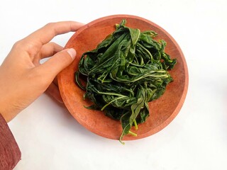 a hand holding a wooden plate filled with Boiled cassava leaves on a white background. healthy and...